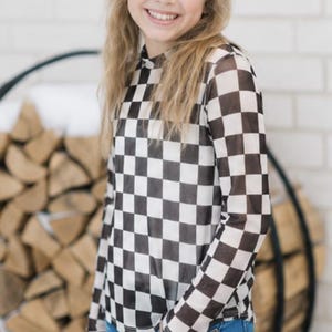 May include: A long-sleeved, black and white checkered shirt. The shirt is paired with blue jeans. The top has a high neckline and a semi-sheer appearance. The model is smiling and has long blonde hair.