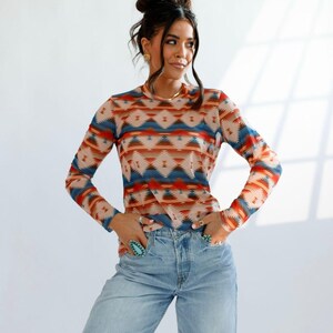 May include: A long-sleeved shirt with a geometric pattern in shades of blue, orange, and beige. The shirt is paired with light-wash, ripped jeans. The top has a crew neck and a fitted silhouette, suitable for casual wear.