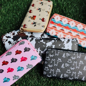 May include: Six different patterned wallets, including a pink wallet with a cowboy hat pattern, a black and white cow print wallet, a black wallet with a white mountain and tree pattern, a cream wallet with a cowboy riding a horse pattern, and a red, blue, and white geometric pattern wallet.