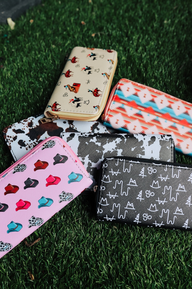 May include: Six different patterned wallets, including a pink wallet with a cowboy hat pattern, a black and white cow print wallet, a black wallet with a white mountain and tree pattern, a cream wallet with a cowboy riding a horse pattern, and a red, blue, and white geometric pattern wallet.