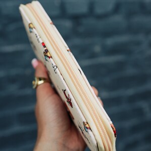 May include: A white zippered wallet with a colorful pattern of people and animals. The wallet is held in a hand with a gold ring and several bracelets.