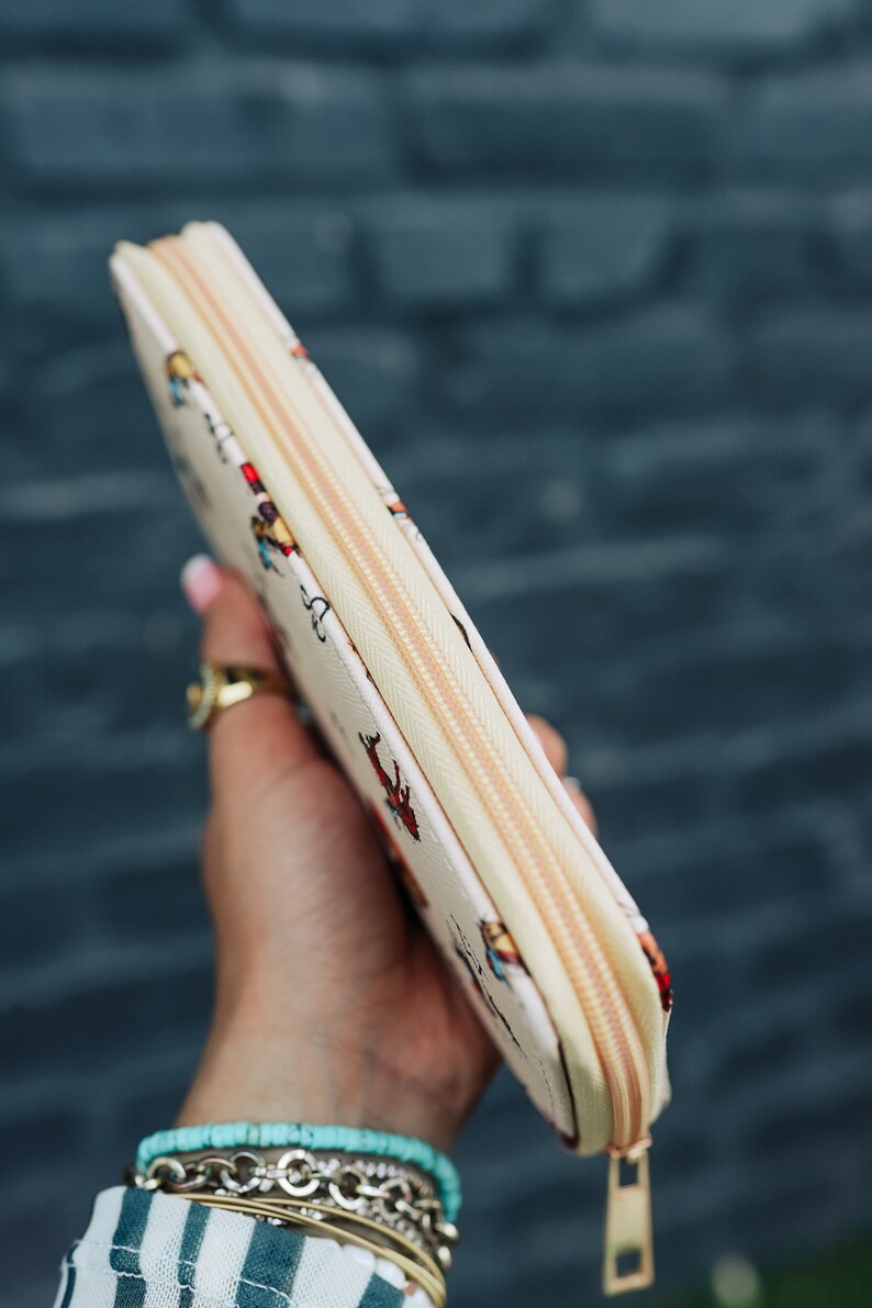 May include: A white zippered wallet with a colorful pattern of people and animals. The wallet is held in a hand with a gold ring and several bracelets.