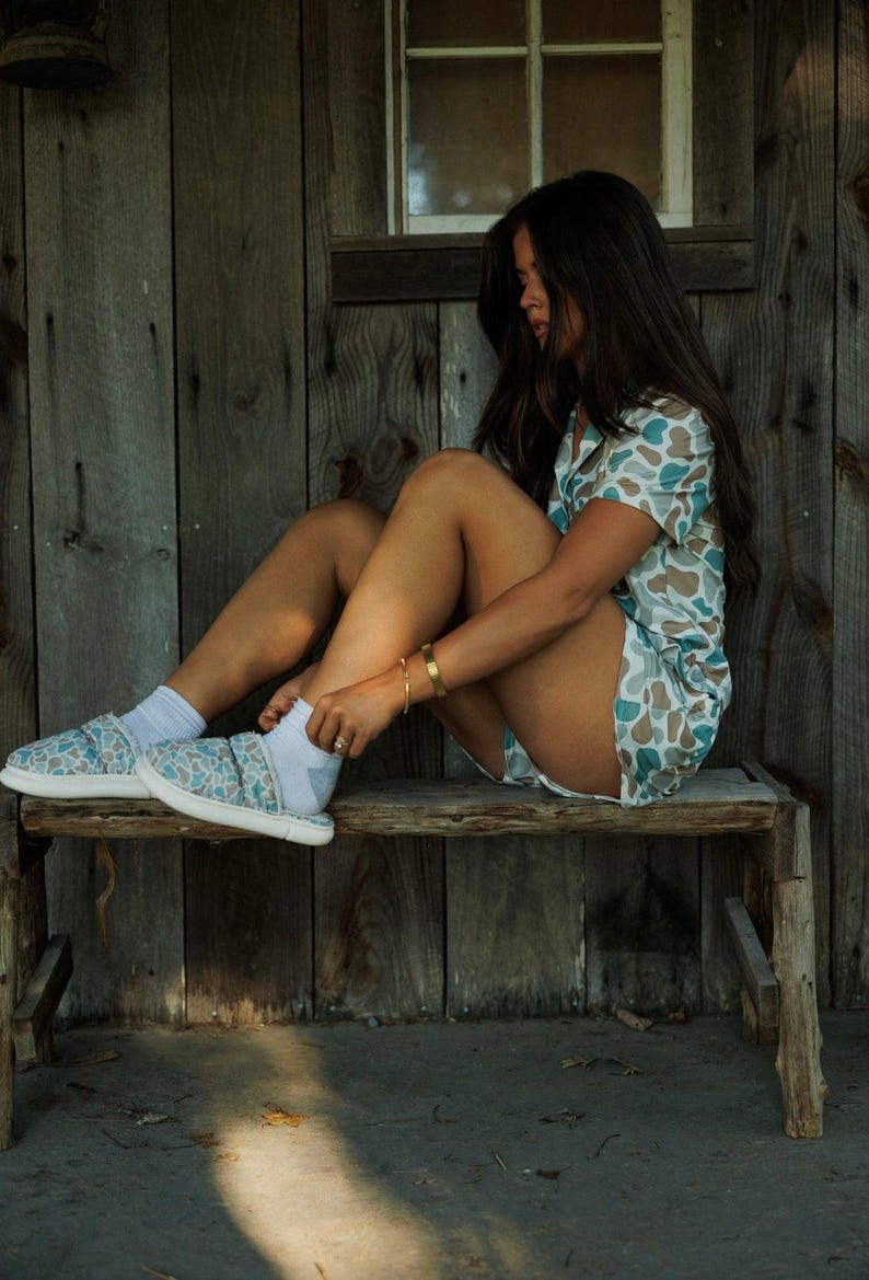 May include: A person wearing a short-sleeved pajama set with a blue and tan camouflage pattern, white socks, and matching slip-on shoes. The person is sitting on a wooden bench. The background is a wooden wall with a window.