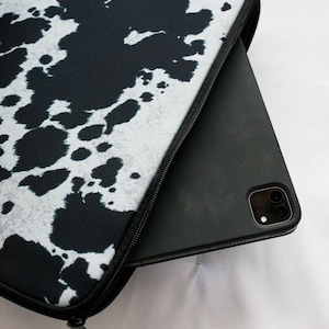May include: A black and white cow print laptop sleeve with a zipper closure. A black tablet is partially inside the sleeve.