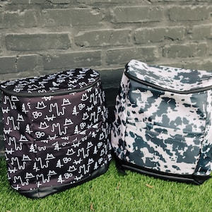 May include: Two black and white patterned backpacks. One backpack has a pattern of mountains, trees, and other symbols. The other backpack has a cow print pattern.