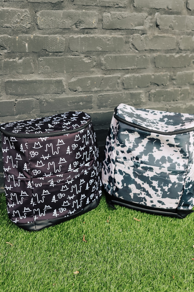 May include: Two black and white patterned backpacks. One backpack has a pattern of mountains, trees, and other symbols. The other backpack has a cow print pattern.
