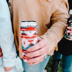 May include: A person is holding a can of Celsius Sparkling Strawberry Watermelon energy drink in a colorful patterned can cooler. The can cooler has a white background with red, orange, blue, and gray geometric patterns.