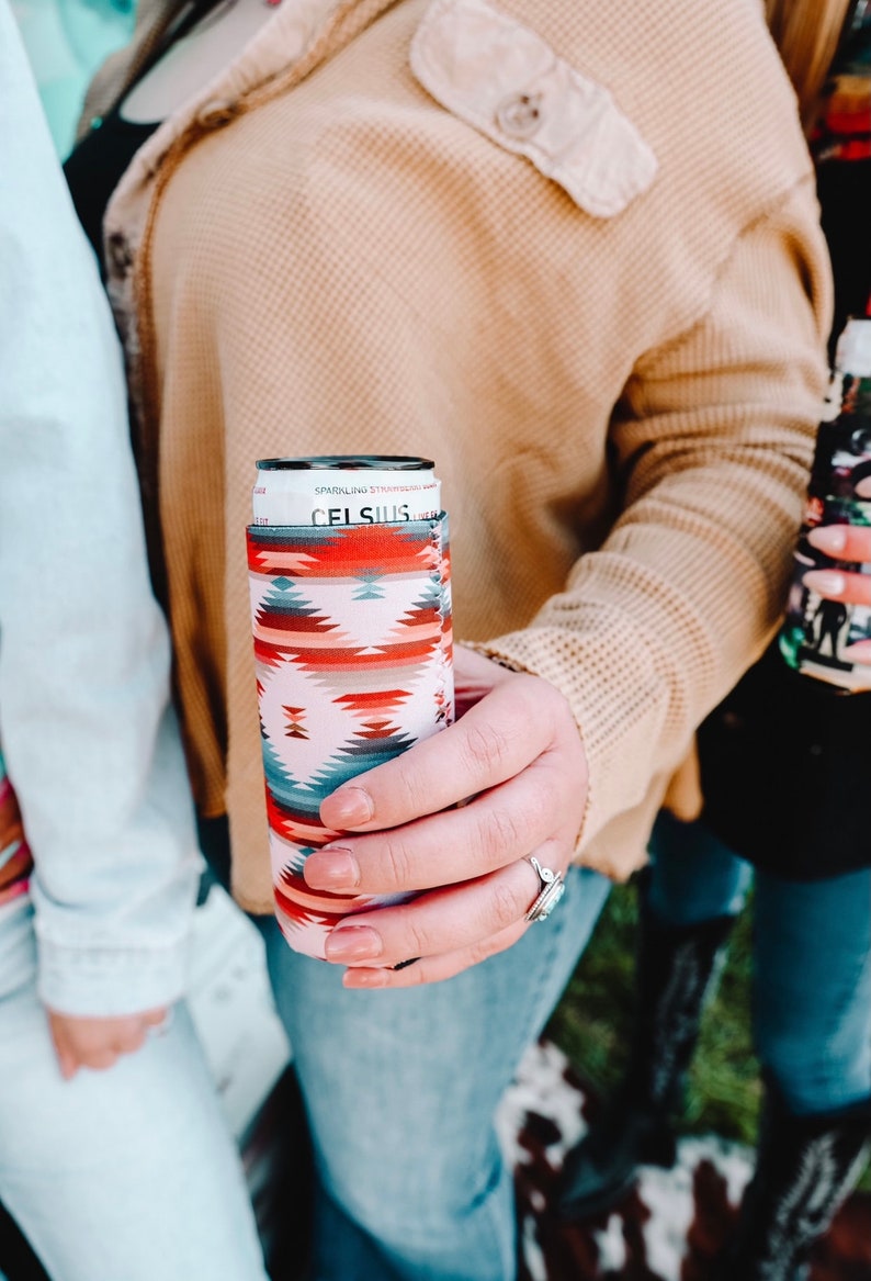 May include: A person is holding a can of Celsius Sparkling Strawberry Watermelon energy drink in a colorful patterned can cooler. The can cooler has a white background with red, orange, blue, and gray geometric patterns.