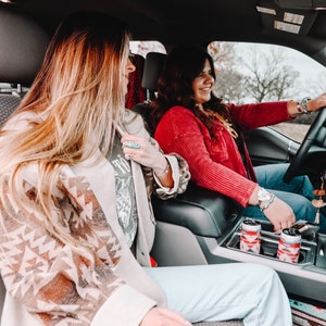 May include: Two women in a car, one wearing a red sweater and the other wearing a brown and white patterned jacket. They are both smiling and looking at the camera. There are two cans of beverage in a cup holder.