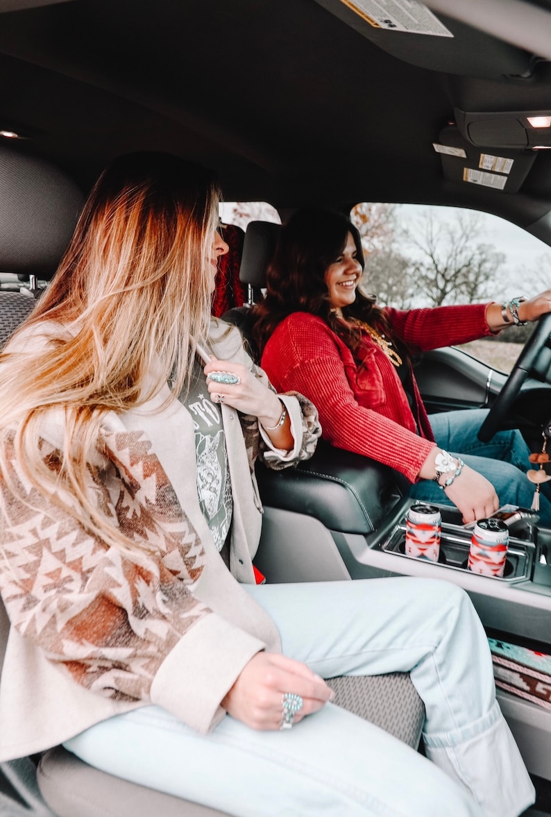 May include: Two women in a car, one wearing a red sweater and the other wearing a brown and white patterned jacket. They are both smiling and looking at the camera. There are two cans of beverage in a cup holder.