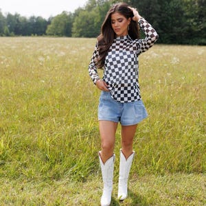 May include: A black and white checkered long-sleeve top, paired with denim shorts and white cowboy boots. The outfit is set against a backdrop of a grassy field and trees, creating a casual, yet stylish look.