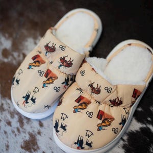 May include: A pair of beige slippers with a repeating pattern of cowboy-themed illustrations. The slippers have a soft, white interior and a white sole. The design includes cowboys, horses, and the number "98".