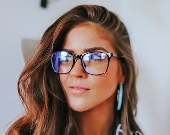 Puede incluir: Una mujer con el pelo largo y castaño que lleva gafas de bloqueo de luz azul con montura negra, una camisa con estampado azul y blanco y pendientes de aro turquesa.