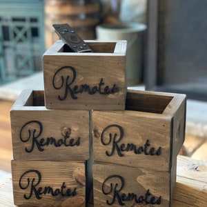 May include: Wooden remote control organizers stacked on a wooden surface. Each box is stained a medium brown and features the word "Remotes" in a decorative font. One box holds a remote control.