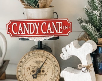 May include: A festive Christmas display featuring a red and white "CANDY CANE LN" sign, a vintage scale, and a white wooden reindeer with a bell. The scale has a cream-colored face and black numbers. Artificial pine and a small pot are also present.