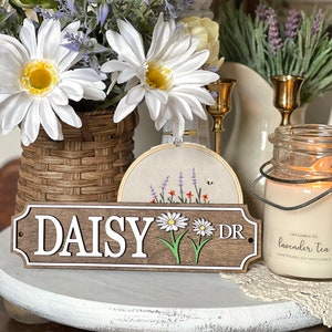 May include: A wooden sign with the text "DAISY DR" in white lettering, featuring daisy flower illustrations. The sign is on a white and gray surface, with a candle and floral arrangement in the background.
