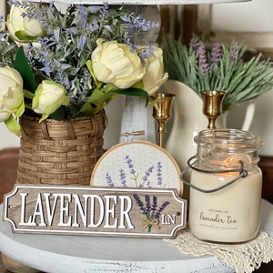 May include: A wooden sign with the word "LAVENDER" in white letters and the initials "LN" in a smaller font. The sign is decorated with a small bouquet of purple lavender flowers. A clear glass jar candle with a white label that reads "LIGH CANDLE CO. lavender tea HAND POURED SOY WAX CANDLE" is in the foreground.