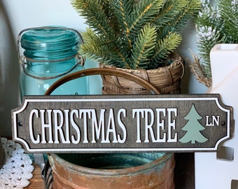 Puede incluir: Un letrero de madera con las palabras "CHRISTMAS TREE" en blanco, con un gráfico de árbol verde y "LN" a la derecha. El letrero tiene un fondo gris oscuro y un borde blanco. El letrero está colocado frente a un recipiente de metal color cobre.