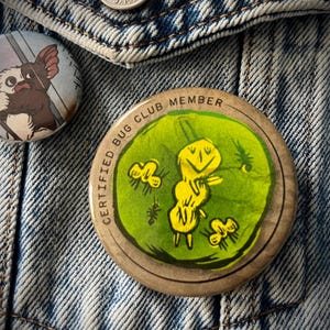 May include: A collection of vintage pins and buttons on a denim jacket. The central pin reads "CERTIFIED BUG CLUB MEMBER" and features a yellow cartoon bug on a green background. Other pins include a cartoon character and a red and yellow pin.