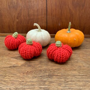 Puede incluir: Cuatro calabazas, dos son de crochet y rojas, una es blanca y otra es naranja. Las calabazas de crochet están sobre una superficie de madera.