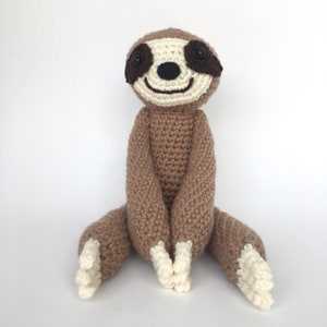 May include: A crocheted sloth plush toy with brown fur, white accents, and black eyes. The sloth is sitting with its arms crossed and its legs outstretched.