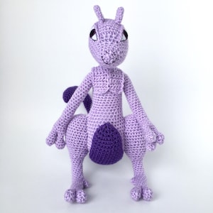 May include: A crocheted purple and lavender kangaroo plush toy with a white background. The toy has a smiling face with black eyes and a purple nose.