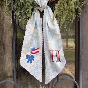 May include: A decorative door scarf with a light blue and white toile pattern, scalloped edges, and a blue bow and American flag embroidery. The letter "H" is embroidered in red.