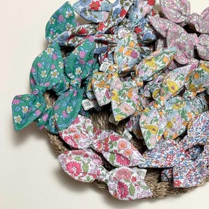 May include: A collection of colorful floral patterned hair scrunchies in a variety of colors, including green, pink, blue, and yellow. The scrunchies are arranged in a circle on a woven surface.