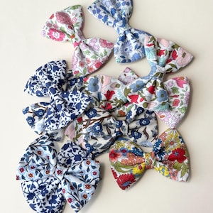 May include: A collection of fabric hair bows in various floral patterns. The bows feature a range of colors, including pink, blue, red, and yellow, with intricate floral designs. The bows are arranged on a light-colored surface.