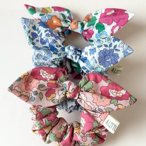 May include: Three floral print scrunchies with bow accents. The scrunchies feature a variety of colorful floral patterns in pink, blue, and orange. Each scrunchie has a small white tag that reads "A LITTLE".