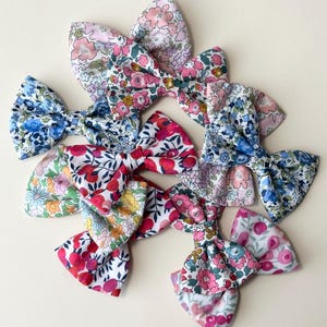 May include: A collection of fabric hair bows featuring various floral patterns in vibrant colours including pink, blue, red, yellow, and green. Each bow is constructed with a central knot and two symmetrical loops, displaying intricate botanical prints.
