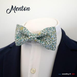 Puede incluir: Una pajarita blanca con un estampado floral verde y amarillo. La pajarita está atada y se muestra sobre una camisa blanca con una chaqueta azul.