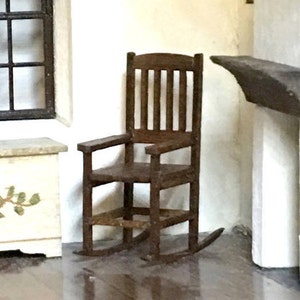 May include: A miniature wooden rocking chair with a dark brown finish. The chair has a slatted back, armrests, and curved rockers. It is placed in a room setting with a window and a small cabinet.