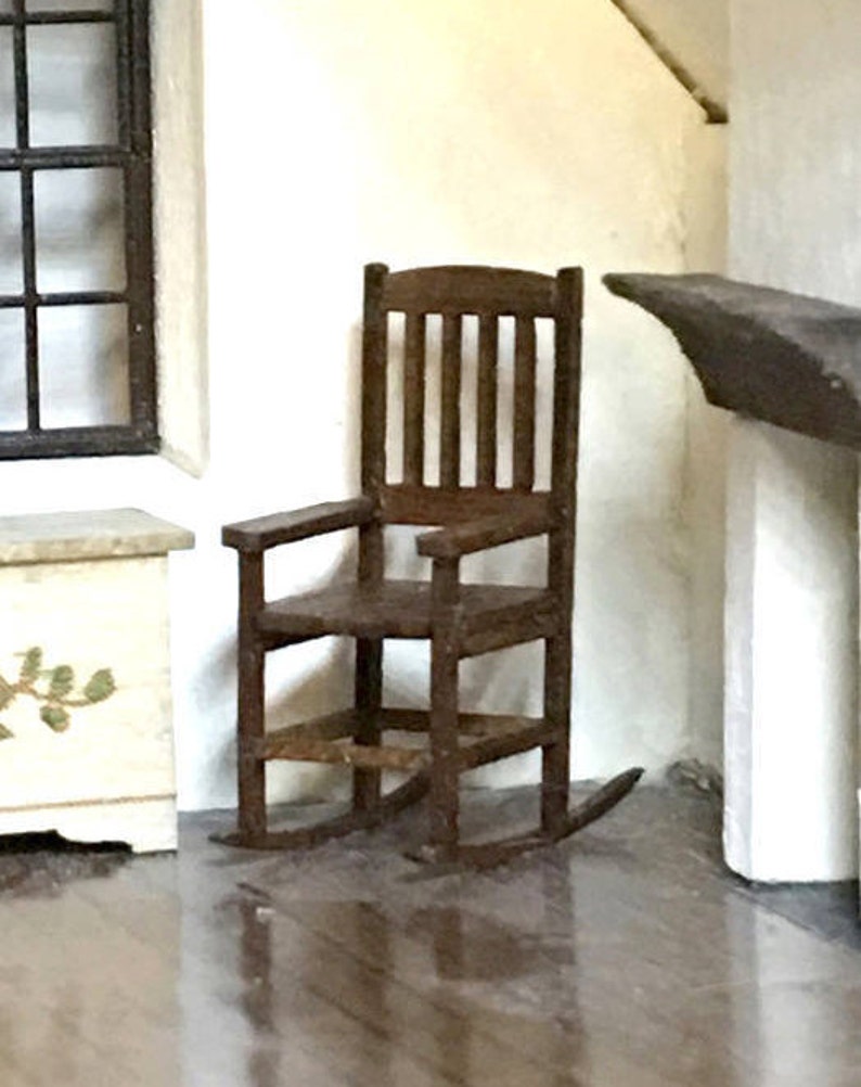 May include: A miniature wooden rocking chair with a dark brown finish. The chair has a slatted back, armrests, and curved rockers. It is placed in a room setting with a window and a small cabinet.