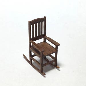 May include: A miniature wooden rocking chair with a dark brown finish. The chair features a slatted back, armrests, and curved rockers. The small-scale furniture piece is set against a white background.