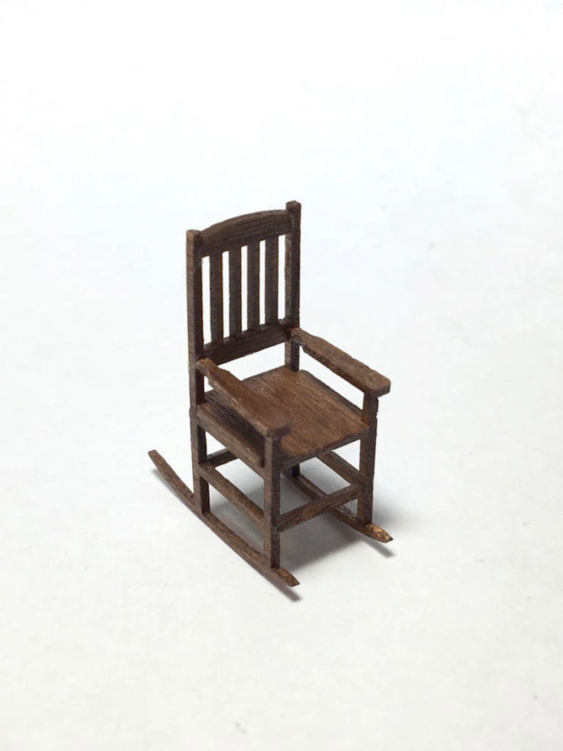 May include: A miniature wooden rocking chair with a dark brown finish. The chair features a slatted back, armrests, and curved rockers. The small-scale furniture piece is set against a white background.