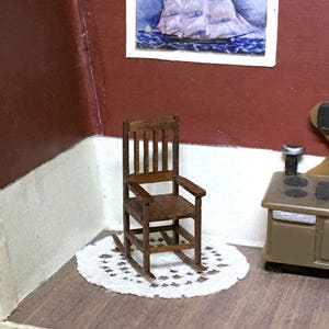 May include: A miniature wooden rocking chair with a dark brown finish sits on a white doily. The chair has a slatted back and armrests. A small painting of a ship hangs on the wall behind the chair.