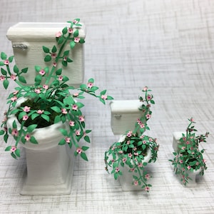 May include: Three white ceramic toilets of different sizes, each with a green leafy plant with pink flowers growing out of the bowl.