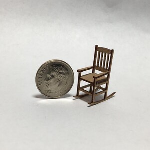 May include: Miniature wooden rocking chair with a light brown finish. The chair has a slatted back and armrests. A U.S. dime is next to the chair for scale. The chair is a small-scale model.