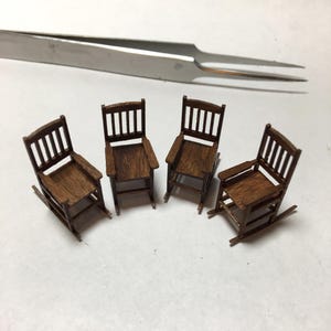 May include: Four miniature wooden rocking chairs with armrests and slatted backs are arranged on a white surface. The chairs are a warm brown color. A pair of silver tweezers is in the background.