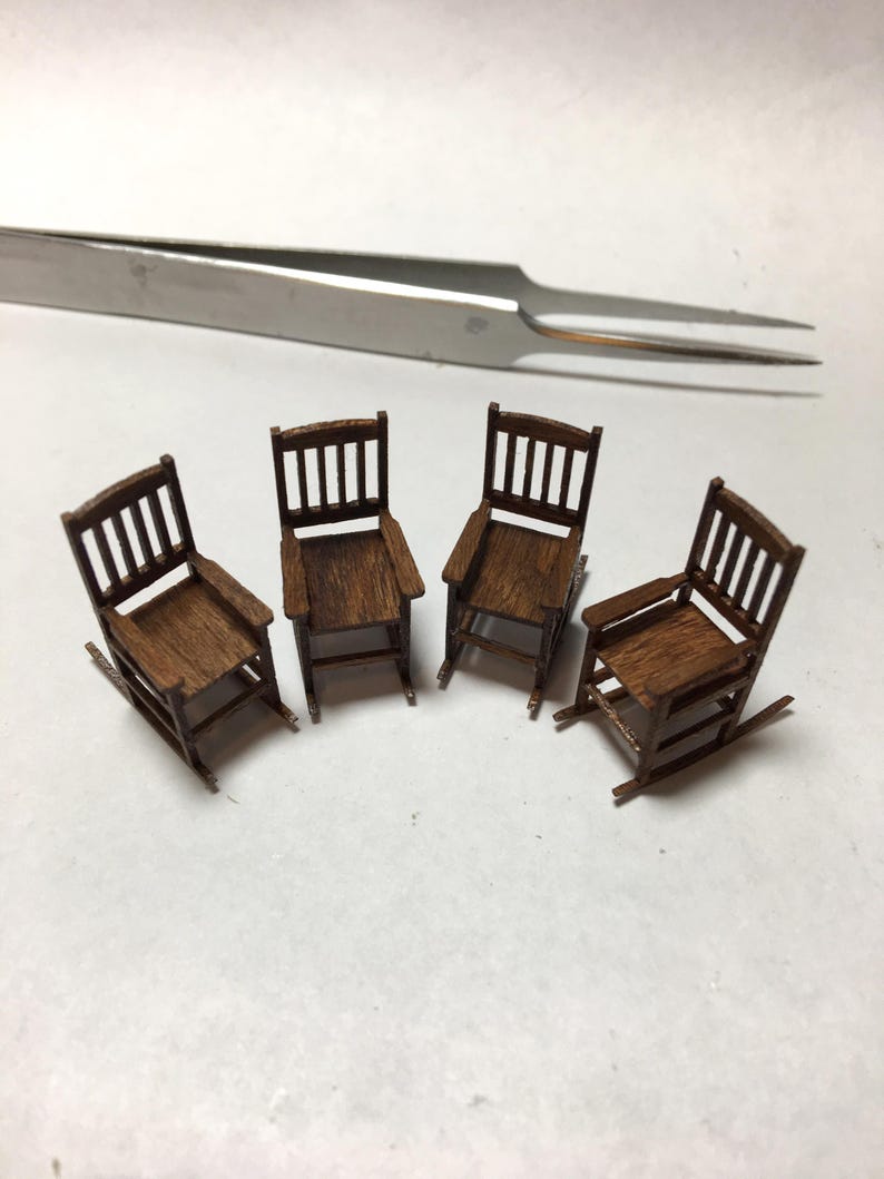 May include: Four miniature wooden rocking chairs with armrests and slatted backs are arranged on a white surface. The chairs are a warm brown color. A pair of silver tweezers is in the background.