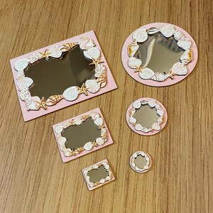 May include: A collection of small decorative mirrors in various shapes: rectangular, square, and round. Each mirror is framed with a pink border and adorned with seashells and starfish. The mirrors are arranged on a wooden surface.