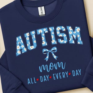 Puede incluir: Sudadera azul marino con la palabra "AUTISMO" en letras grandes de color azul claro llenas de margaritas blancas. Debajo, un lazo azul claro y la palabra "mamá" en cursiva. La frase "TODO EL DÍA TODOS LOS DÍAS" está debajo.