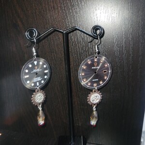 May include: Black and silver earrings featuring watch faces with the words "Rolex" and "Relox". Each earring has a decorative silver charm and a dangling faceted crystal bead. The earrings are displayed on a black metal stand.
