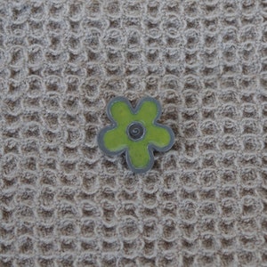 May include: A green flower-shaped pin with a silver swirl in the center. The pin is on a beige textured background.