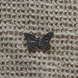 May include: A detailed, silver-colored butterfly pin. The butterfly has intricate wing patterns and is set against a textured, beige background. The pin is a decorative accessory, suitable for clothing or other fabric items.