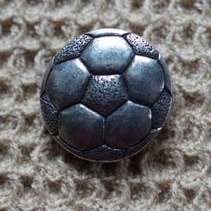 May include: A silver-toned metal button shaped like a soccer ball. The button has a textured surface and a raised design of the soccer ball's panels.