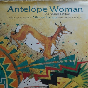 May include: Book cover with an illustration of an antelope running across a colorful geometric pattern. The title of the book is "Antelope Woman: An Apache Folktale" and the author is Michael Lacapa.
