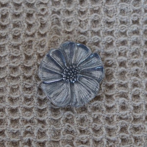 May include: A silver-colored flower-shaped brooch with detailed petal and center designs. The brooch is set against a textured, tan background.