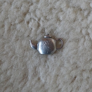 May include: A small, silver-colored teapot charm with a rose design on the front. The charm has a handle, spout, and a lid. The teapot is set against a textured, off-white background.